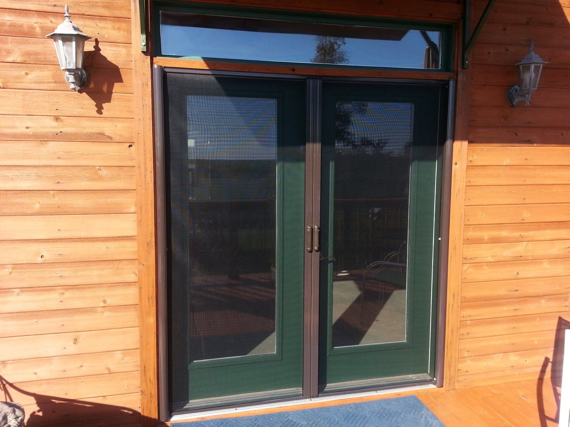 Green double doors with screen panels, flanked by lights on a wood-paneled building.