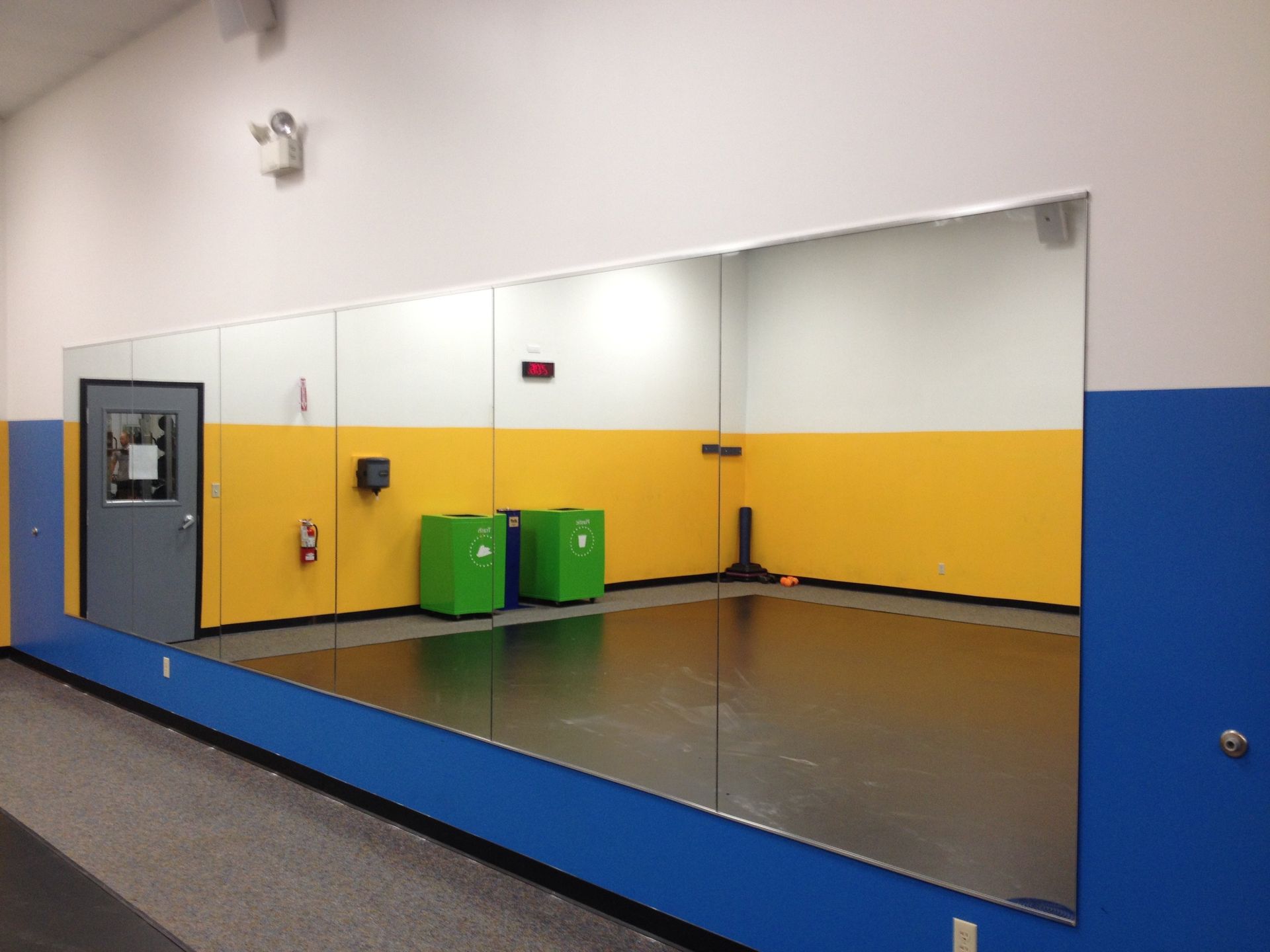 Gym interior with large wall mirrors, yellow and blue walls, and green storage containers.