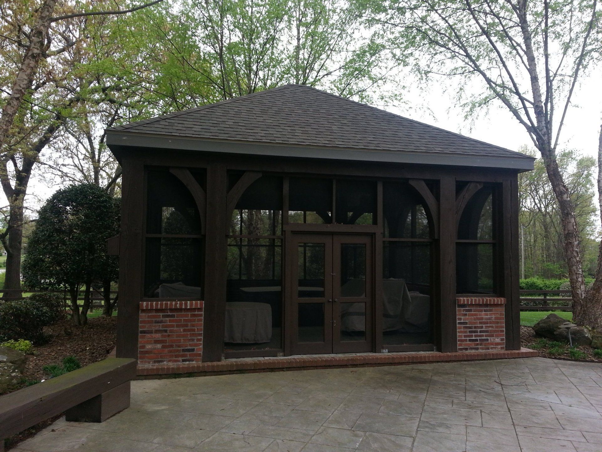Dark brown screened gazebo with a brick base on a gray patio. Trees in the background.