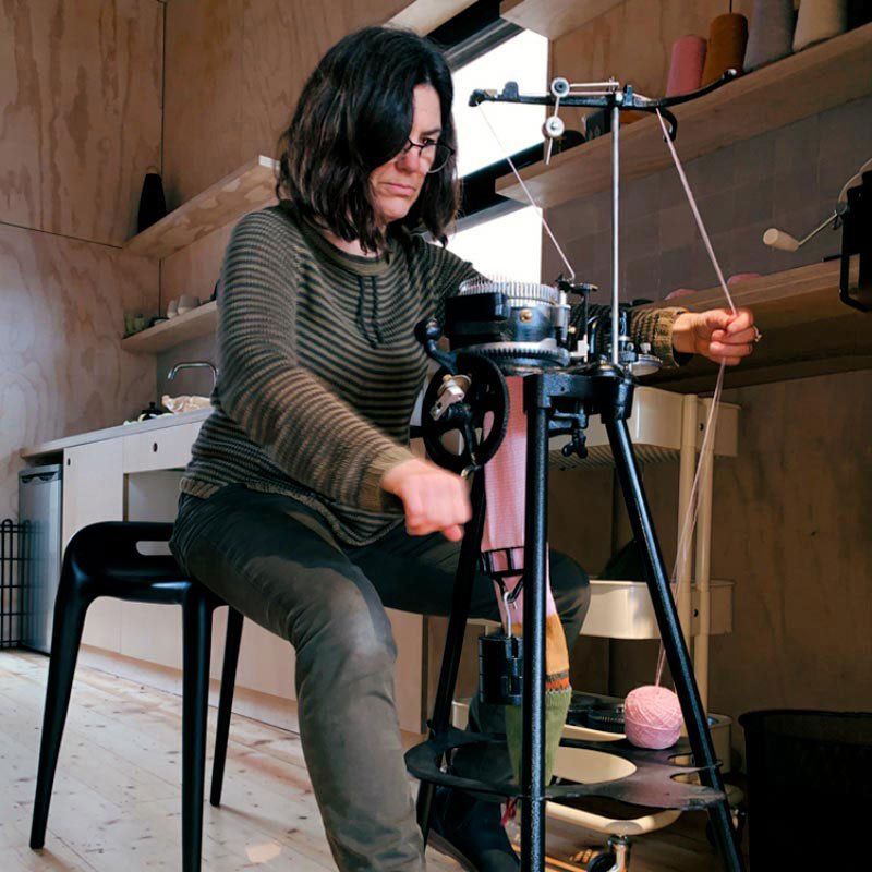 Linda Castles sits behind a mechanical circular sock knitting machine, turning the handle on the machine in order to operate it