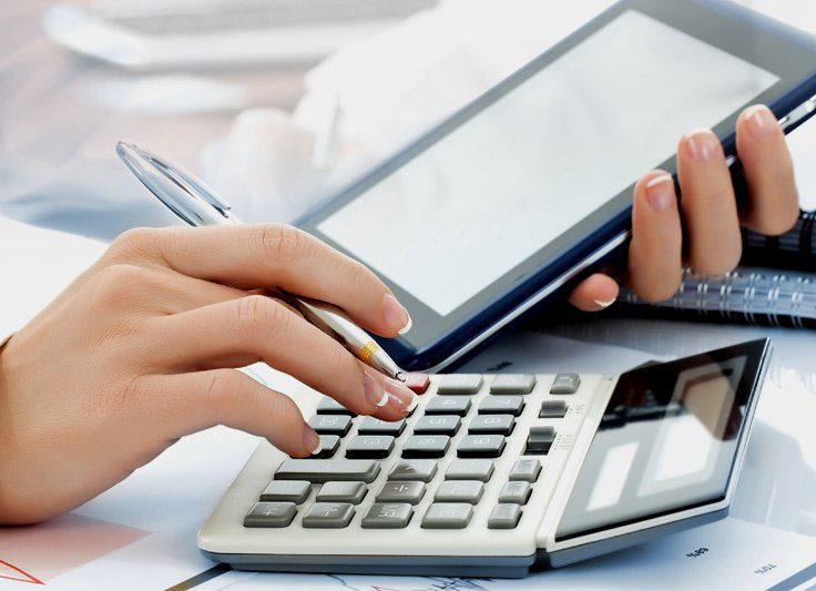 Person's hands using a calculator and holding a pen, with a tablet in the background.