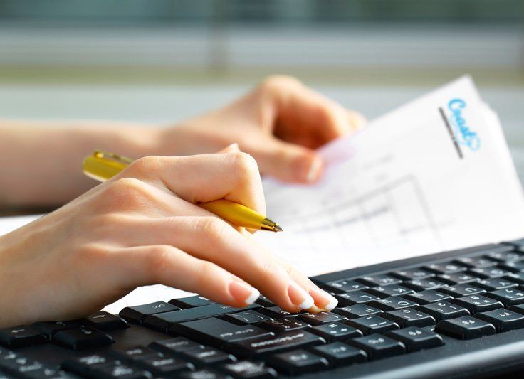 Hands typing on keyboard, holding paper with logo. Pen rests on the paper.