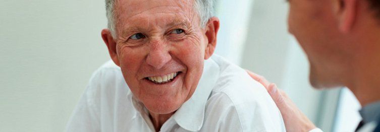 Older man smiling as a person's hand rests on his shoulder; doctor's arm in the foreground.