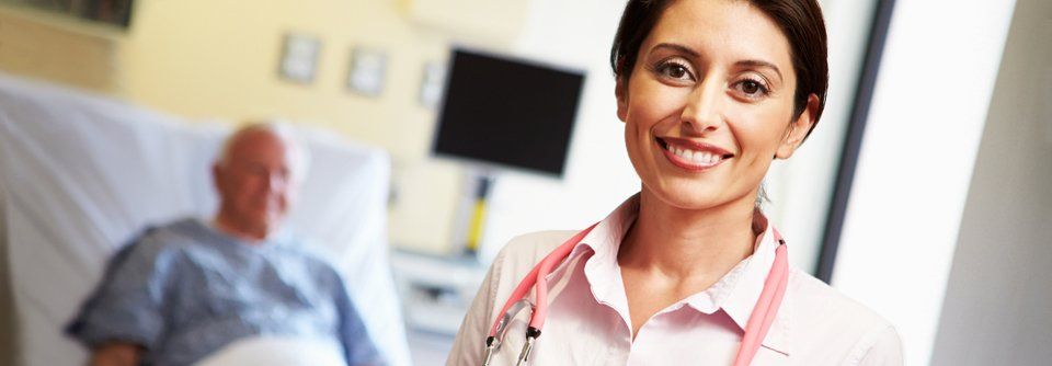 Smiling healthcare worker with stethoscope, patient in bed blurred in the background. Hospital room.