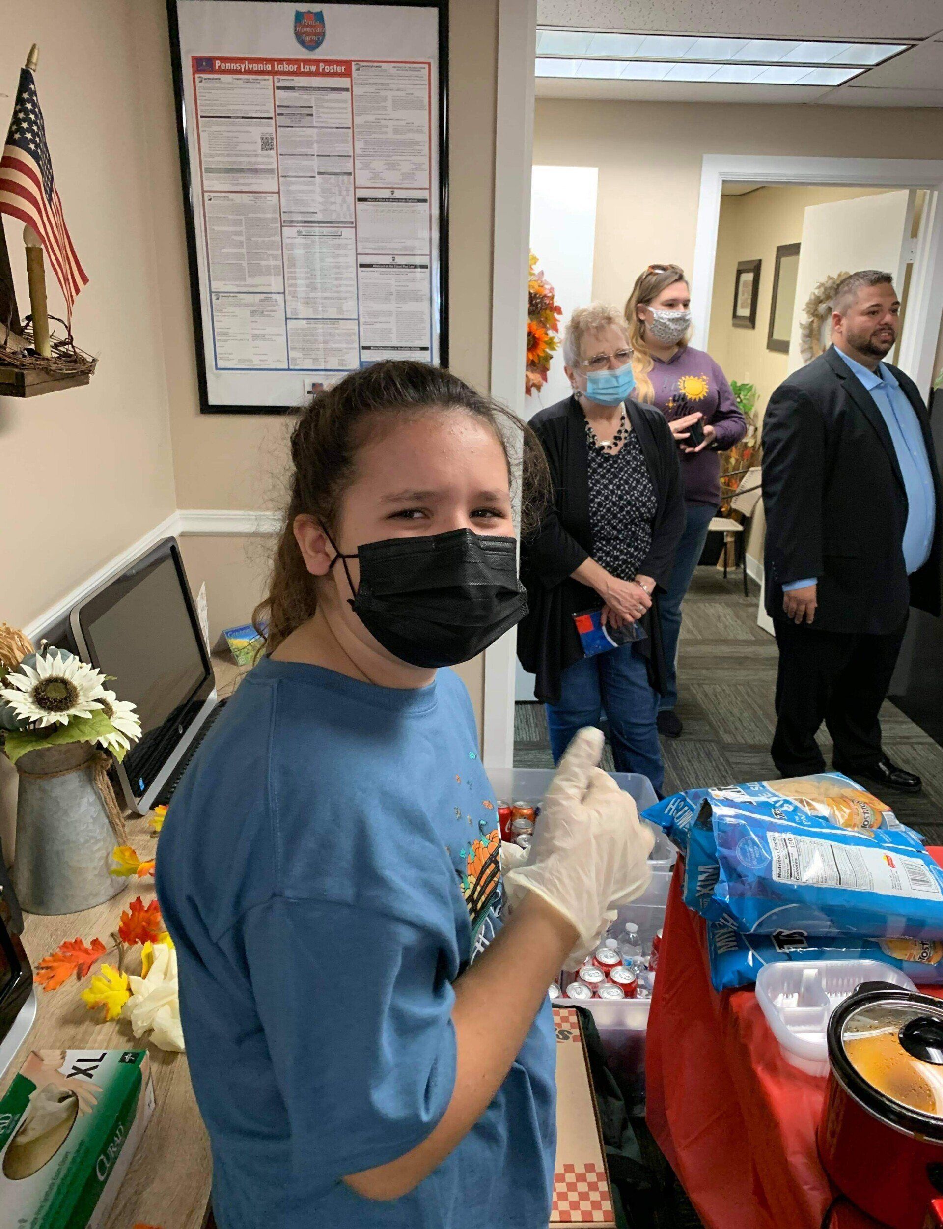 Person in blue shirt and mask, pointing while holding a glove, at a table with snacks, with others in background.