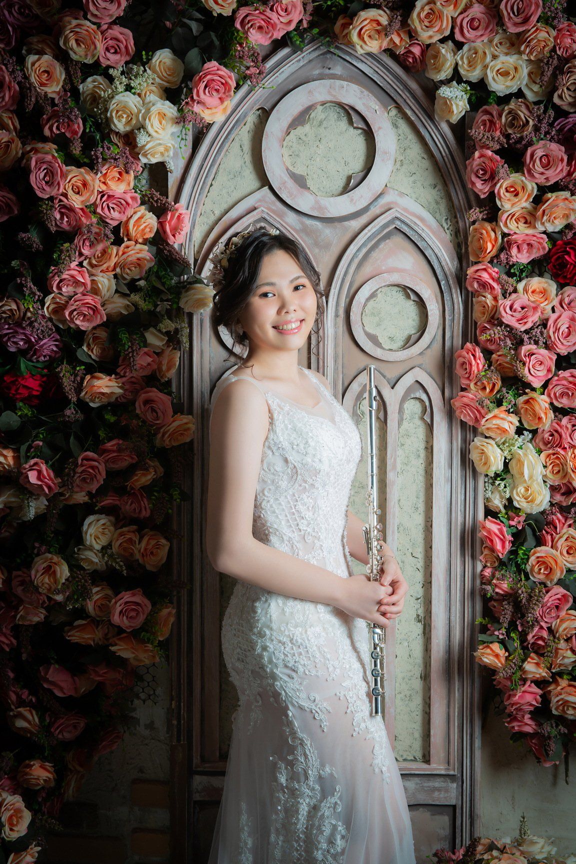 一位身穿白色蕾絲長裙的女子在花牆和拱形窗戶前擺姿勢拍照。
