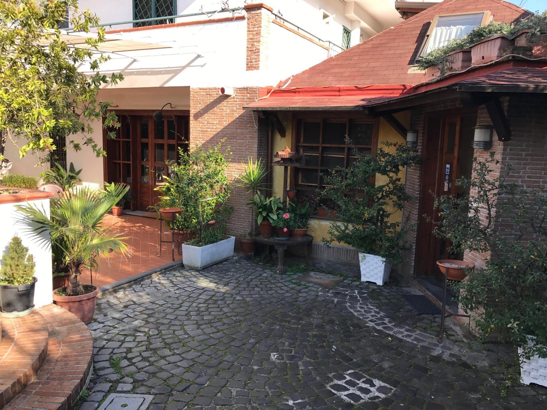 Ingresso nel cortile con pavimentazione in pietra e piante in vaso, che conduce a porte e edifici in legno.