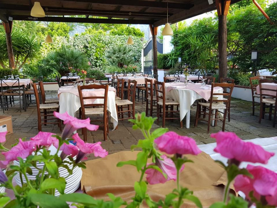 Ristorante all'aperto con tavoli apparecchiati con tovaglie bianche, dettagli rosa e circondato da una vegetazione lussureggiante.