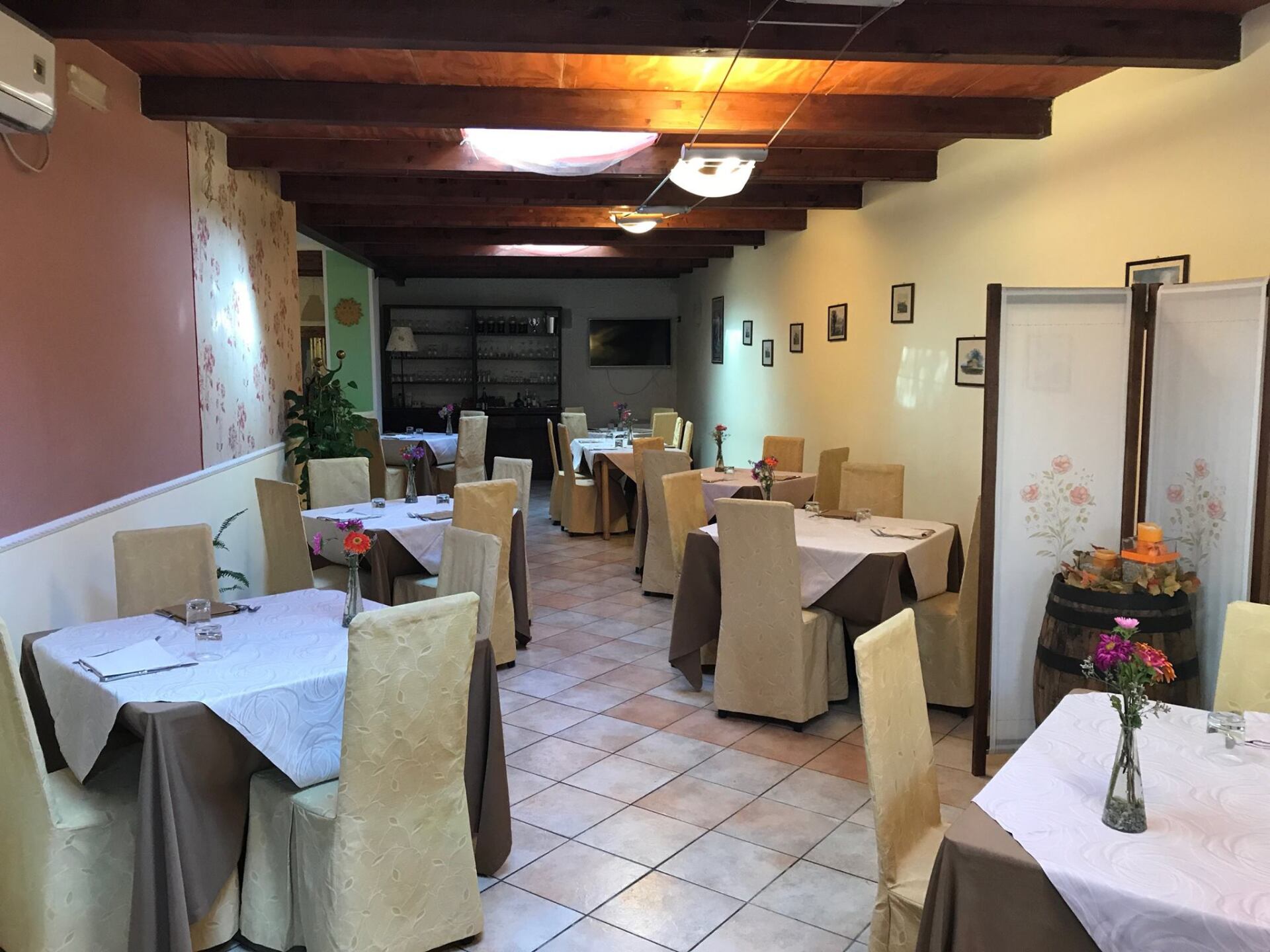 Interno del ristorante con tavoli apparecchiati per cenare; pareti beige, dettagli marroni e travi in legno.