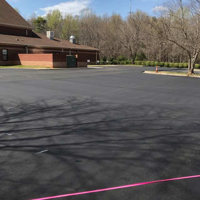 Large, newly paved parking lot in front of a brick building and trees, with a pink line on the asphalt.