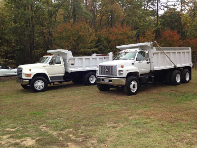 Two white dump trucks parked on grass, autumn trees in background.