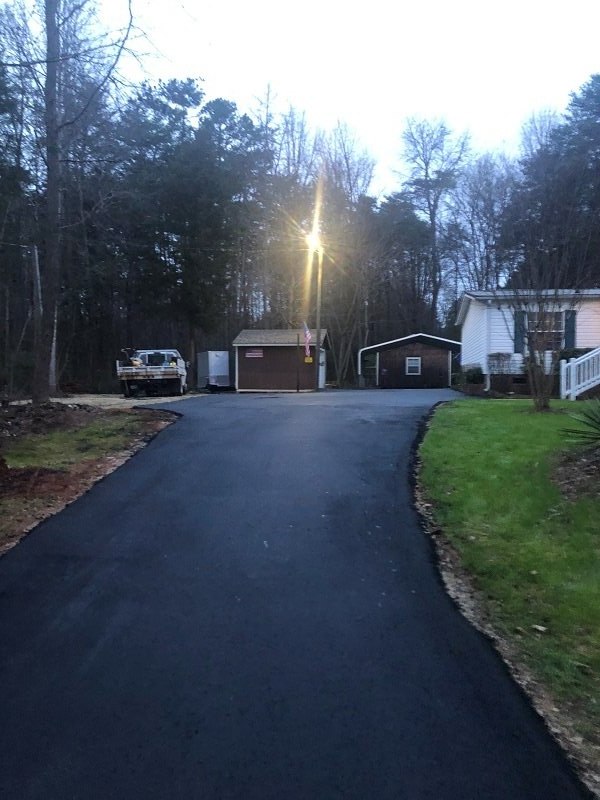 Newly paved asphalt driveway leading to a group of buildings and parked vehicle surrounded by trees.