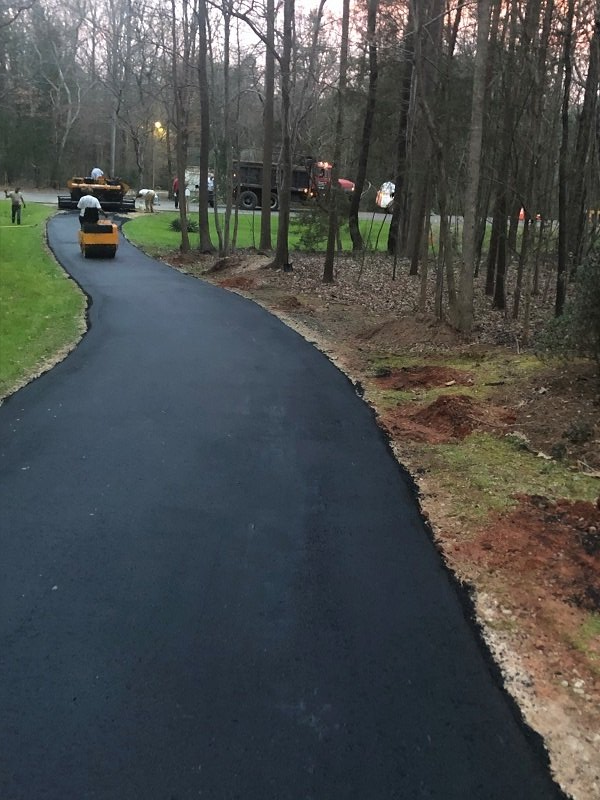 Paved black asphalt driveway being rolled by a machine with trees and grass bordering.