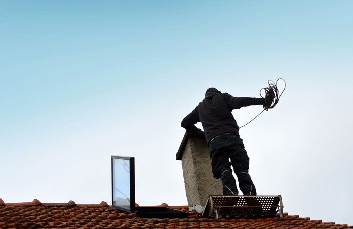 Worker on a red-tiled roof adjusting a chimney with a cable against a clear sky