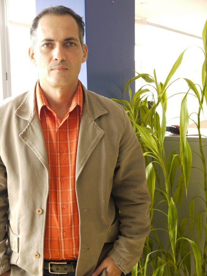 Un hombre vestido con una chaqueta y una camisa naranja está parado frente a una planta.