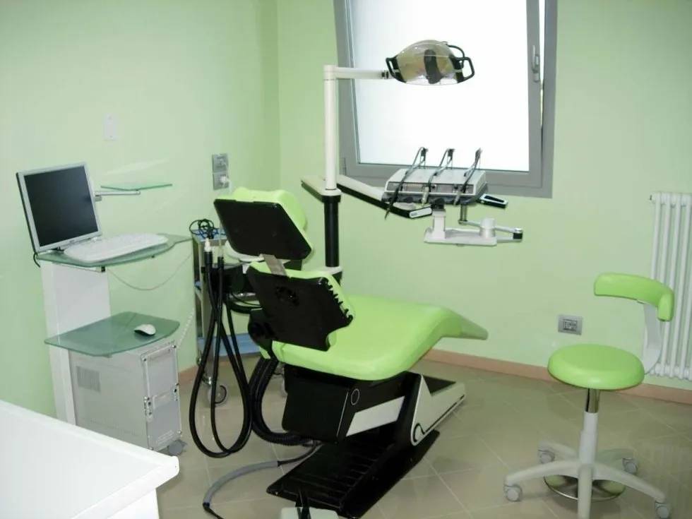 Sala visite di uno studio dentistico con sedia verde, lampada a soffitto, computer e sgabello.