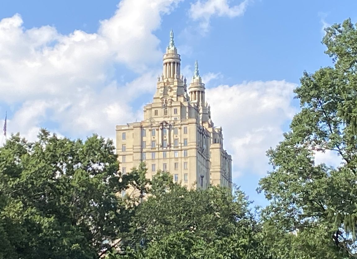 Tall ornate building rises above green trees against a cloudy blue sky.