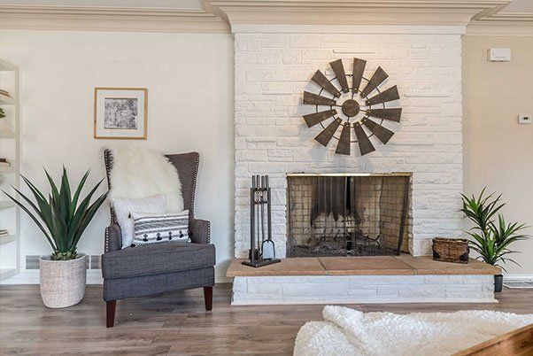 A living room with a fireplace , chair , plants and a windmill on the wall.