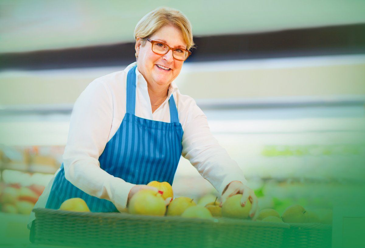 A woman in a blue apron is standing next to a basket of apples in a grocery store.