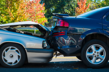Two cars with severe rear-end collision damage on a road, blue car hitting a silver car in daylight.