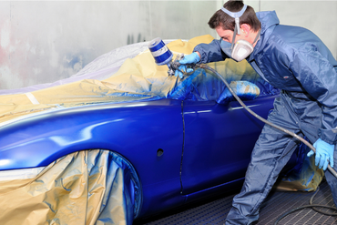 Person spray-painting a blue car in a workshop, with masked surfaces taped off for painting