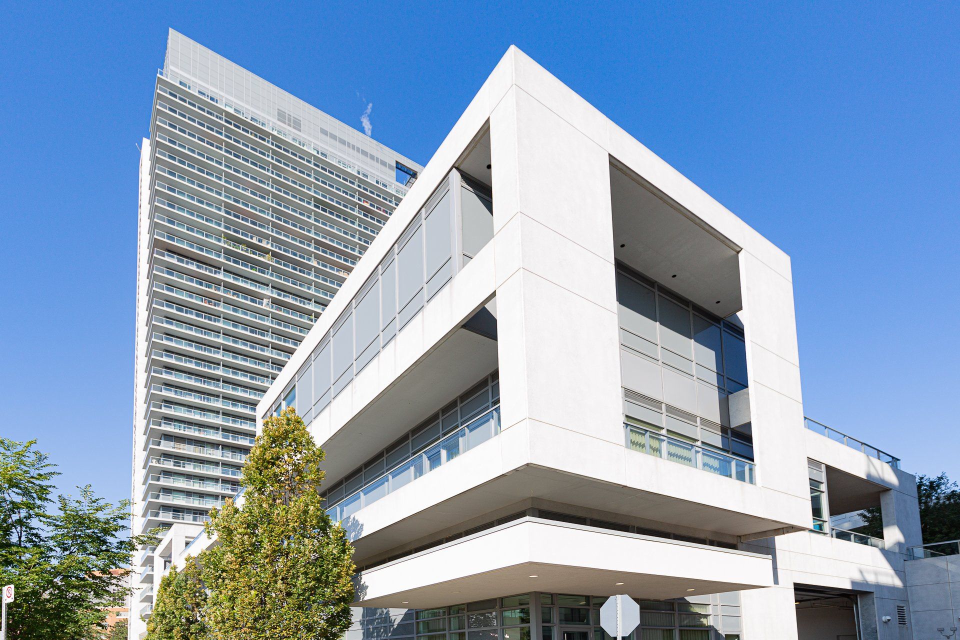 A modern, white, multi-level building with large windows beside a tall, grid-patterned skyscraper under a clear blue sky.