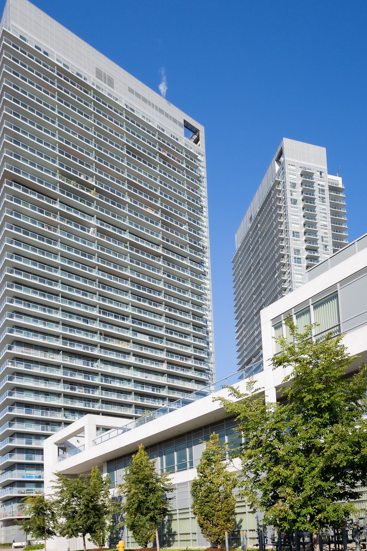 Two modern high-rise apartment buildings tower above a lower-level commercial building and small trees against a blue sky.
