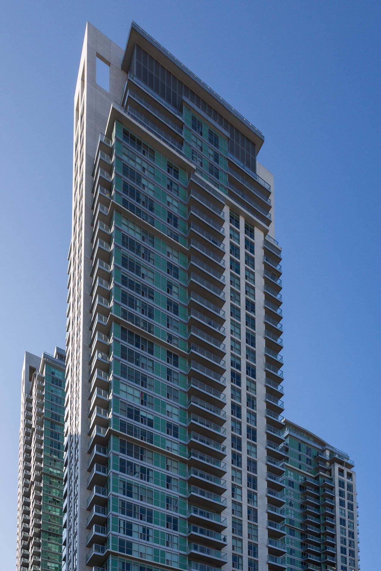 A tall, modern residential skyscraper with turquoise glass windows and multiple balconies under a clear blue sky.