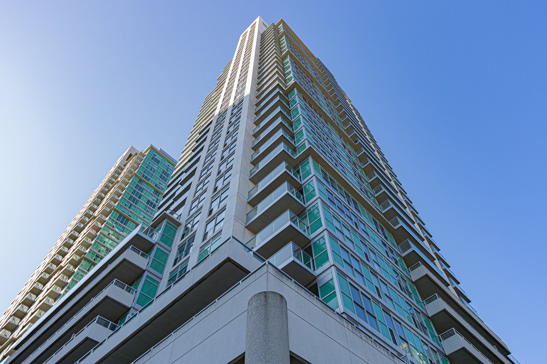Low-angle view of two modern residential high-rise towers with glass balconies against a clear blue sky.