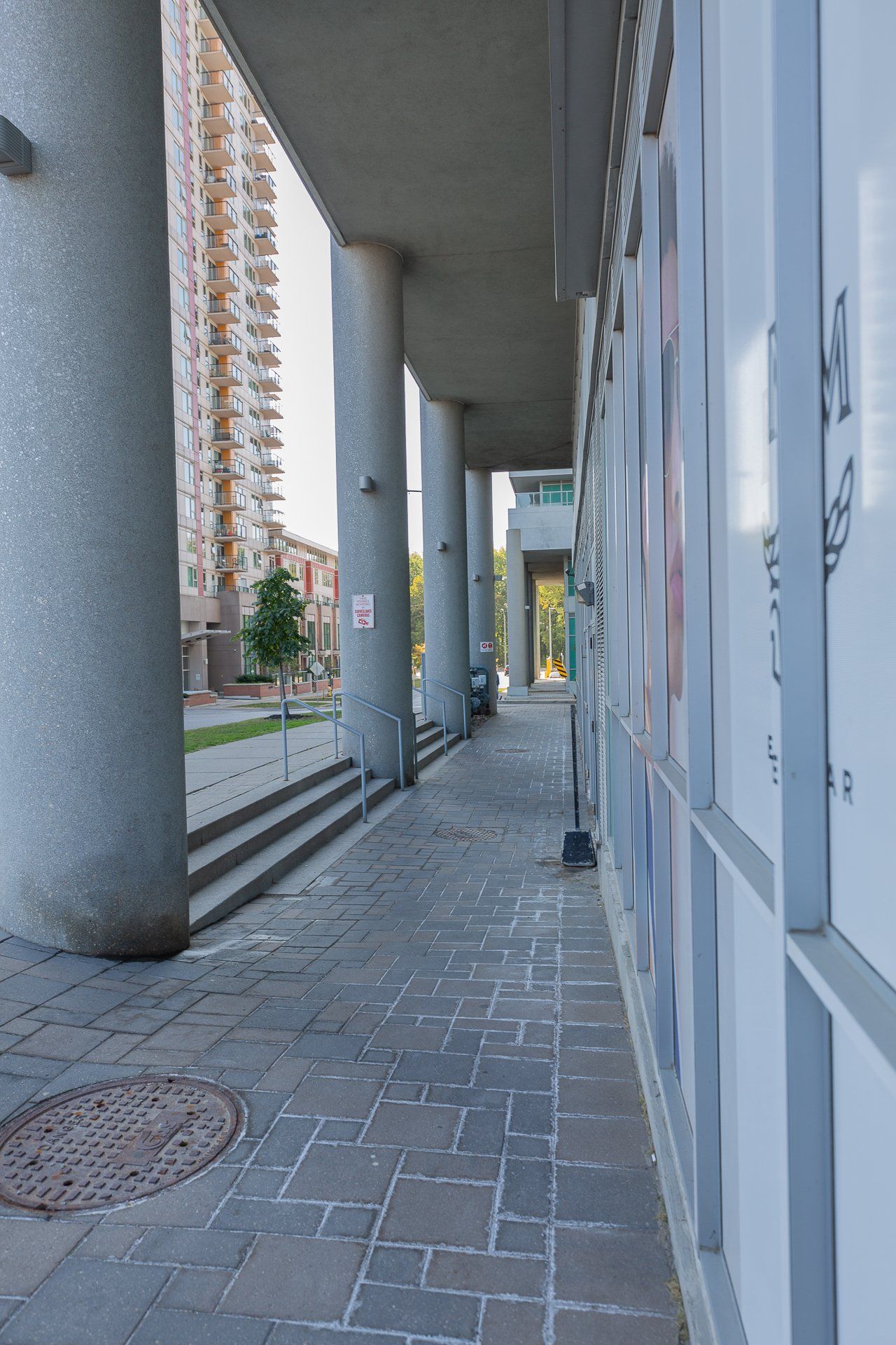 An outdoor covered walkway lined with gray pillars leads past a glass storefront with posters, alongside a stone sidewalk.
