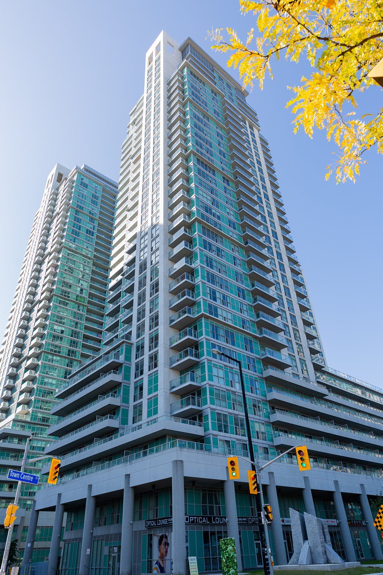 Two tall, modern glass residential towers rise against a bright blue sky, with autumn leaves visible in the corner.