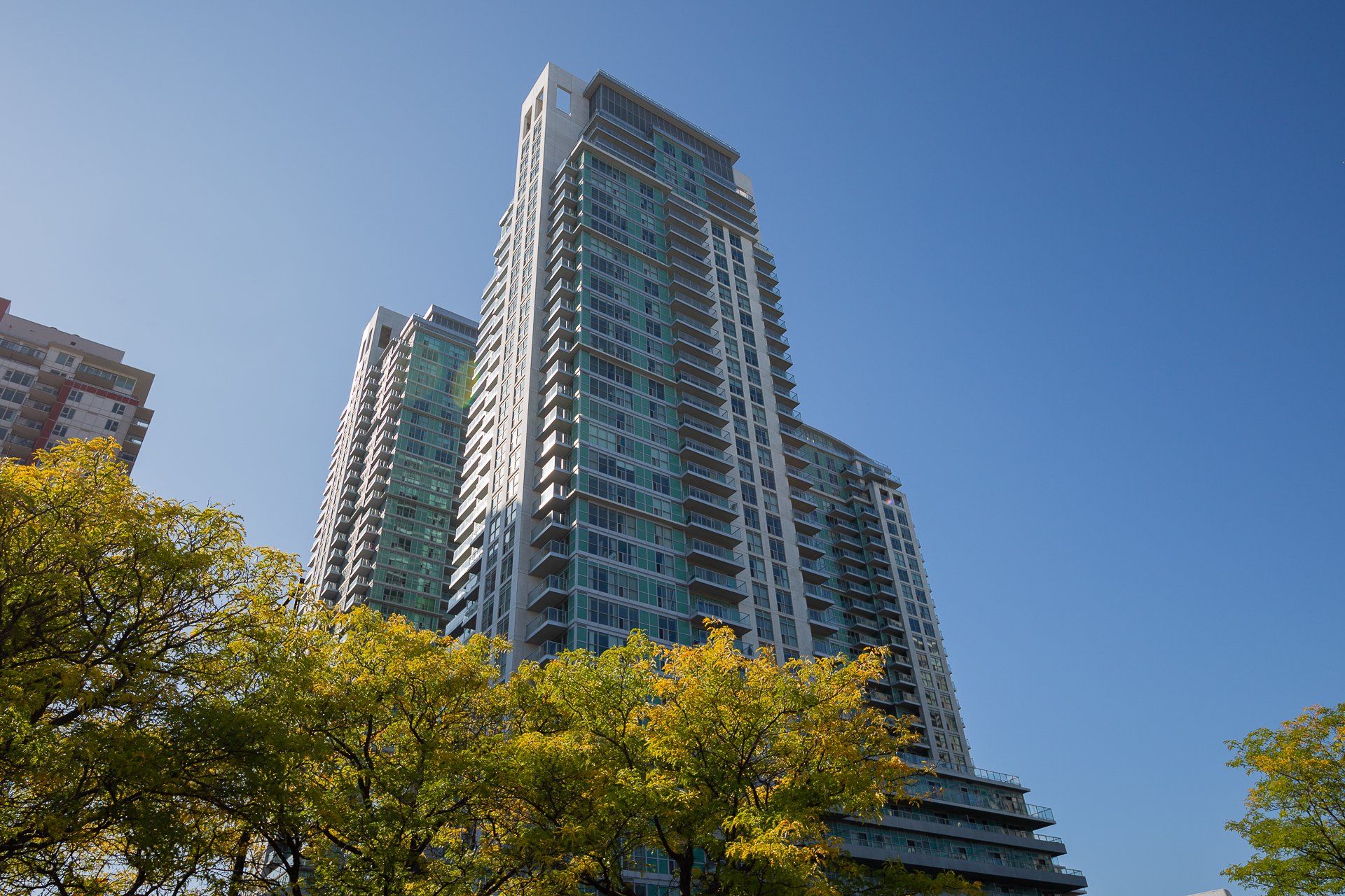 A tall, modern residential skyscraper with blue-tinted glass and balconies, framed by trees with yellow autumn leaves.