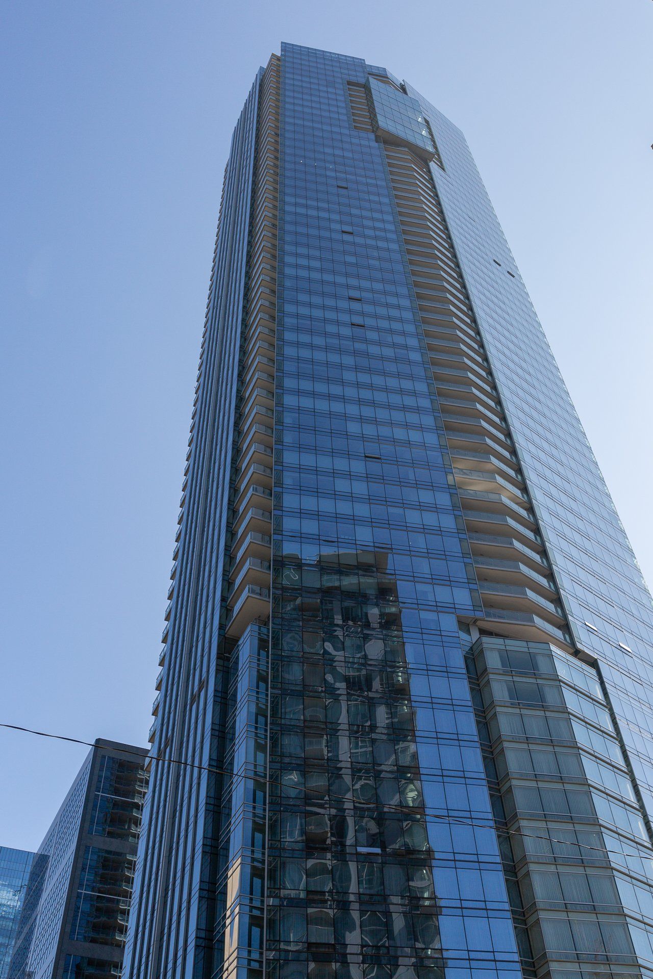 A towering, modern glass skyscraper reflects the bright blue sky against a clear, sunny backdrop.