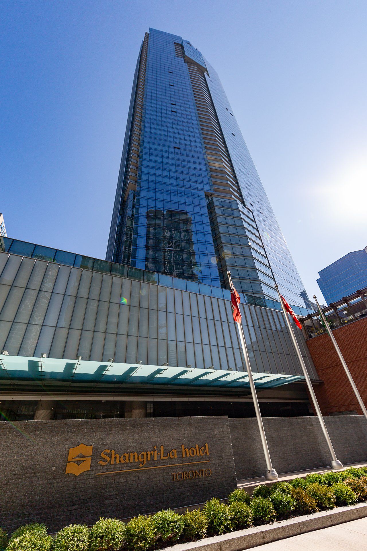 The tall, glass-facade Shangri-La hotel skyscraper stands against a clear blue sky, seen from a low street-level angle.