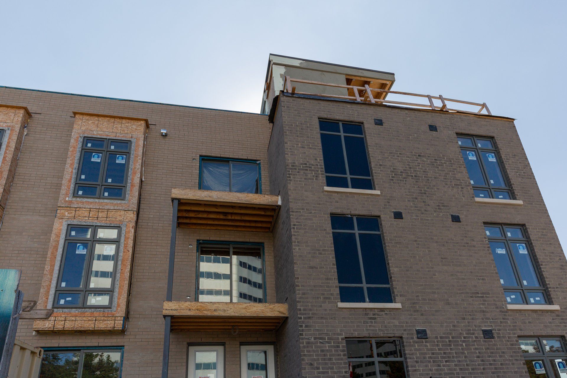 A modern apartment building under construction, featuring gray brick facade, wooden balcony framing, and installed windows.
