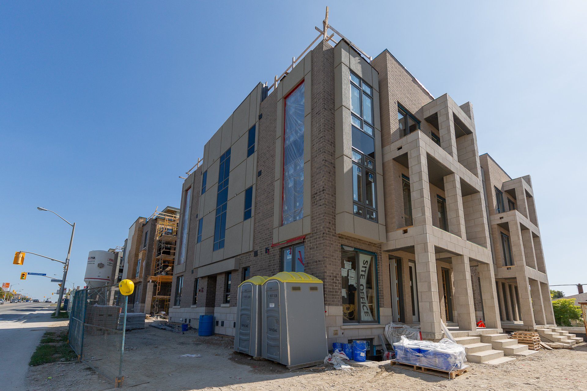 A multi-story building under construction with beige brick siding, large glass windows, and two portable toilets in front.