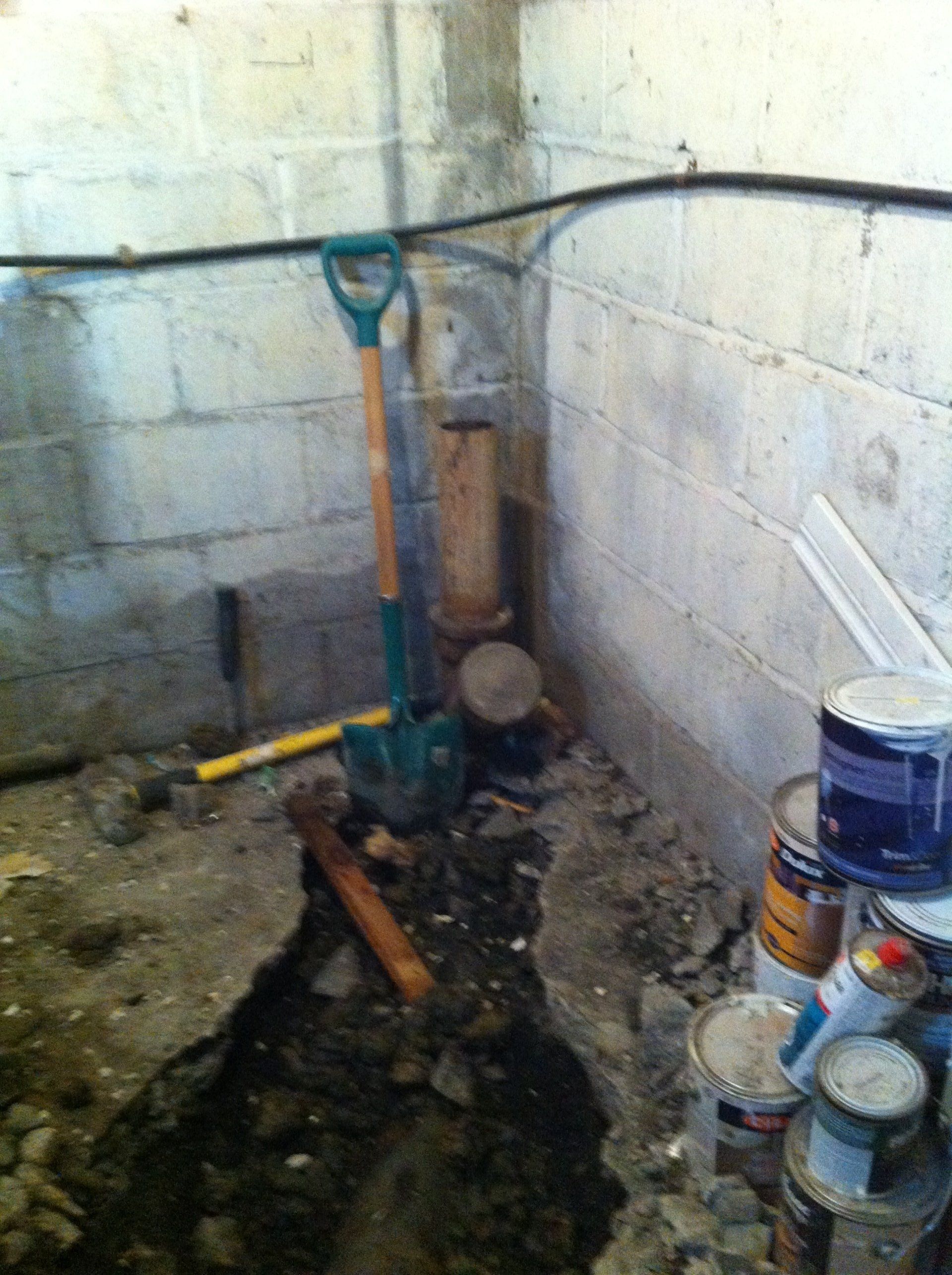 A shovel stands in a basement corner near a broken concrete floor, a vertical pipe, and several paint cans.
