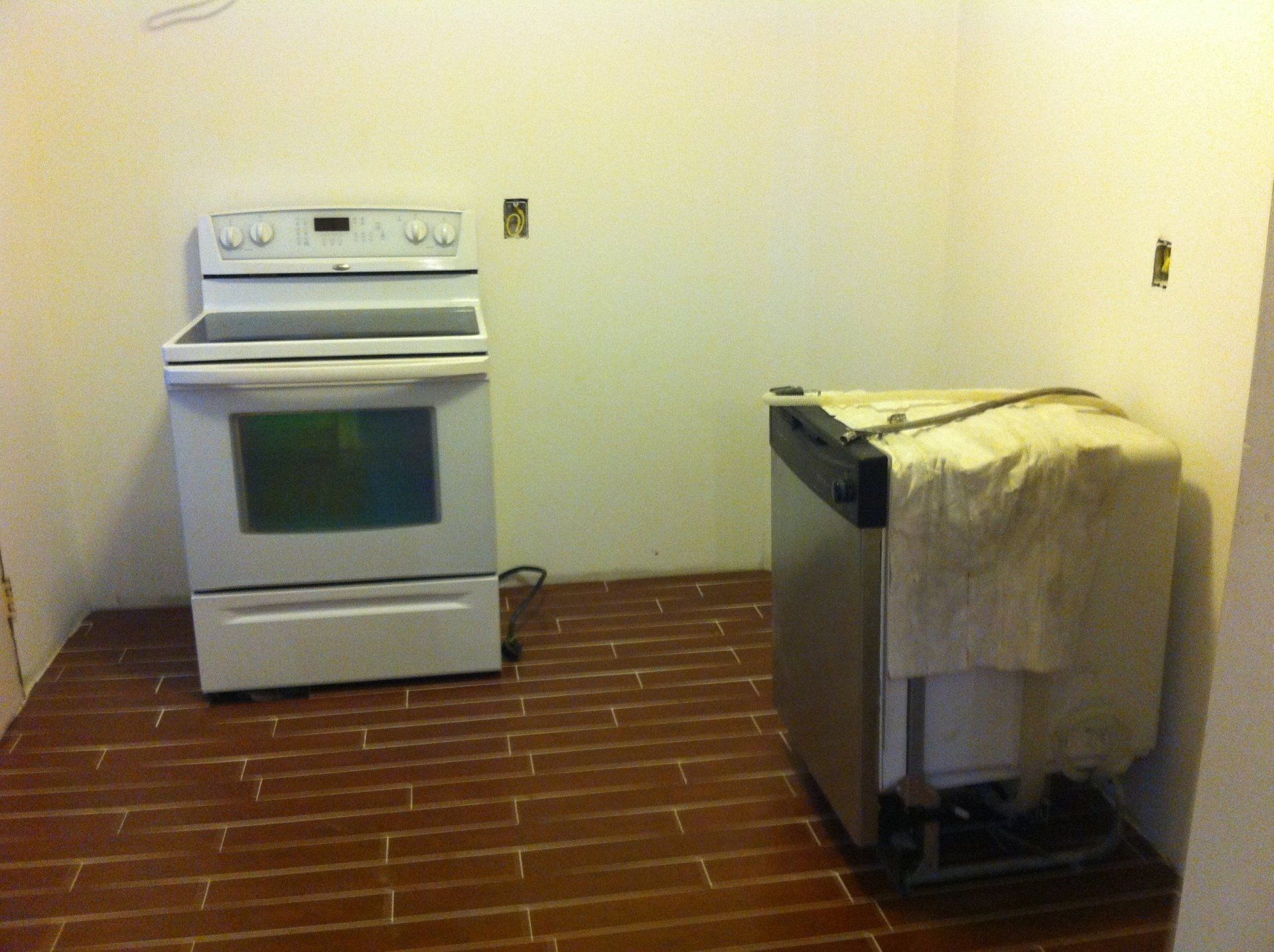 A white stove stands next to a stainless steel dishwasher covered by a cloth in a room with a red-brown tiled floor.