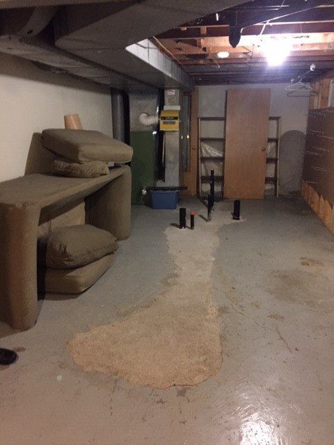 A cluttered basement with gray floors, a long table, tan cushions, shelving, and black pipes protruding from the floor.