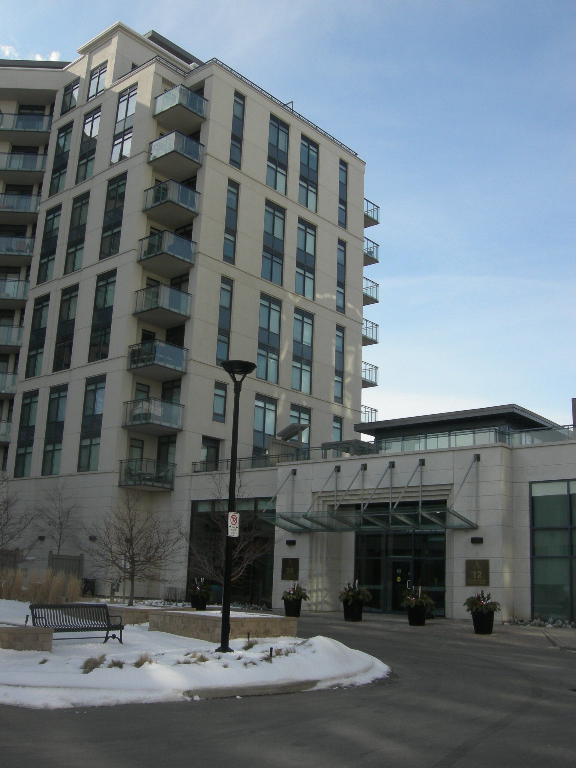 A tall, light-colored apartment building with balconies and a glass-canopied entrance, surrounded by snow on a sunny day.