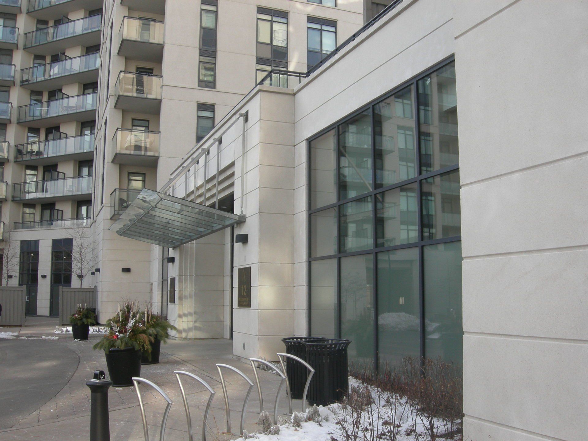 A modern apartment building entrance with a glass awning, bike racks, and potted plants on a concrete sidewalk.