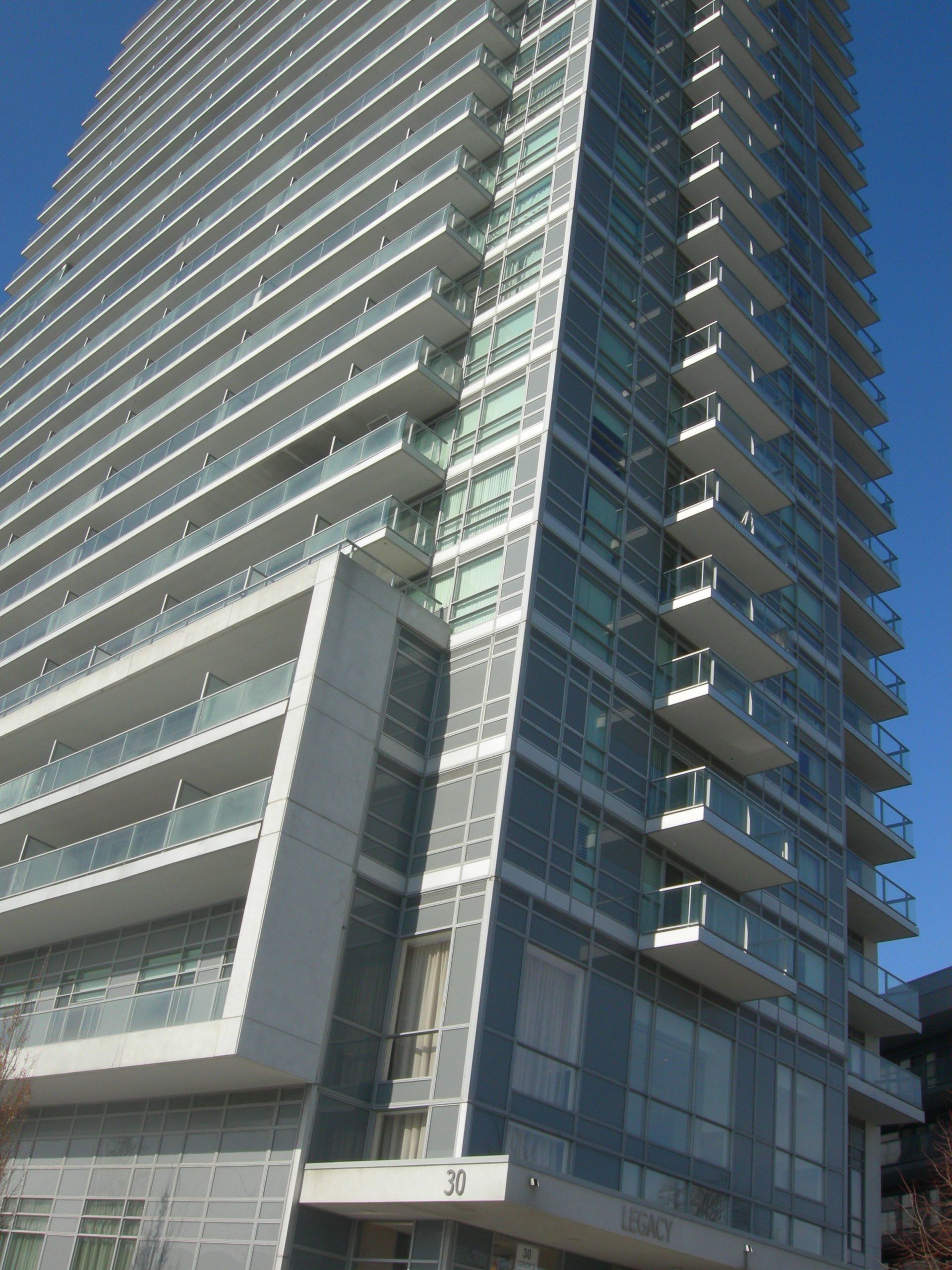 A tall, modern residential skyscraper with glass balconies, light gray concrete accents, and a clear blue sky background.
