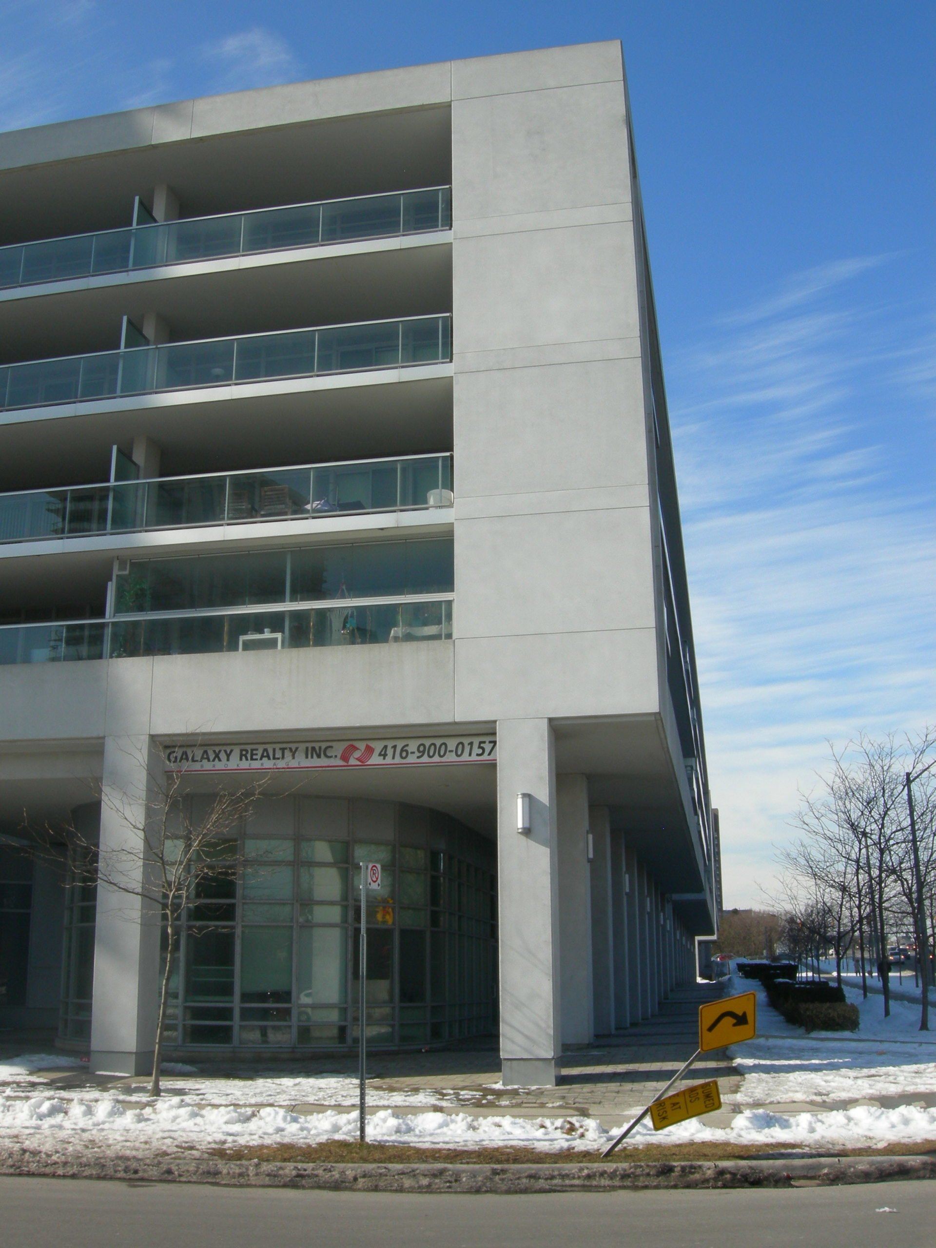 A modern gray concrete building with multiple balconies over a glass-walled ground level, set on a snowy street corner.