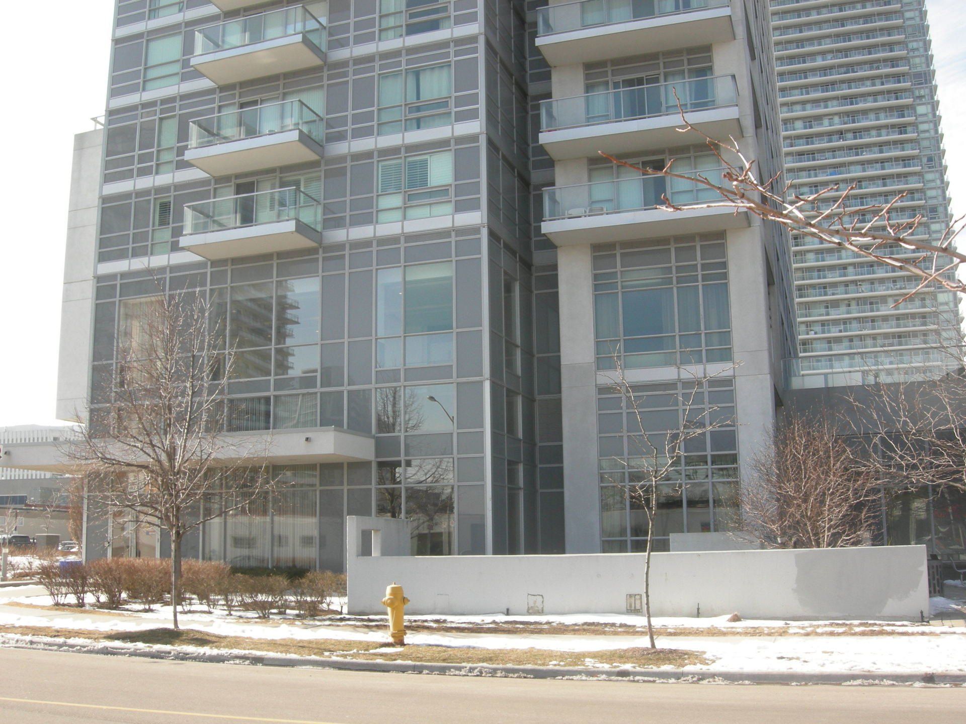 A modern, multi-story glass apartment building with balconies, surrounded by bare trees and snow-covered ground.