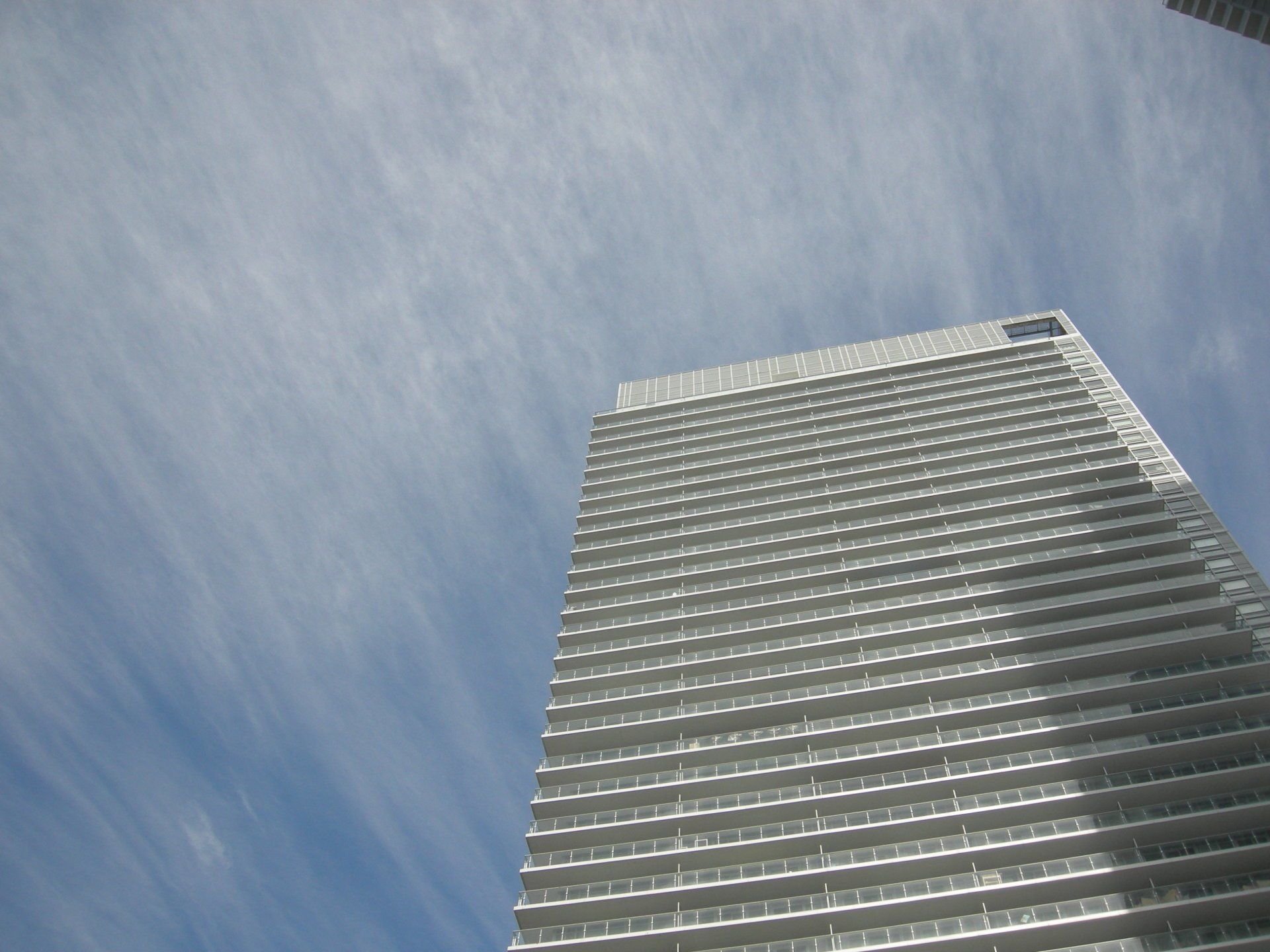 A low-angle view of a tall, modern white skyscraper reaching toward a bright blue sky with wispy clouds.