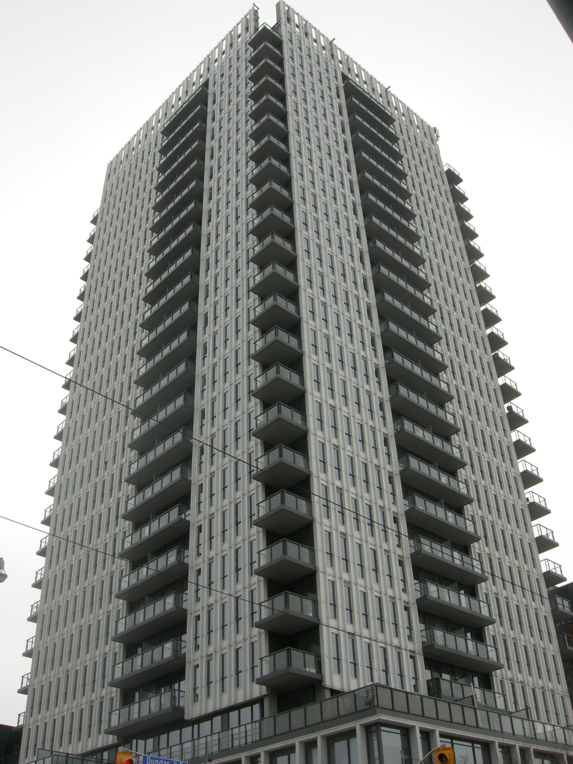 A modern, tall apartment building with a white textured facade and stacked balconies on a cloudy day.