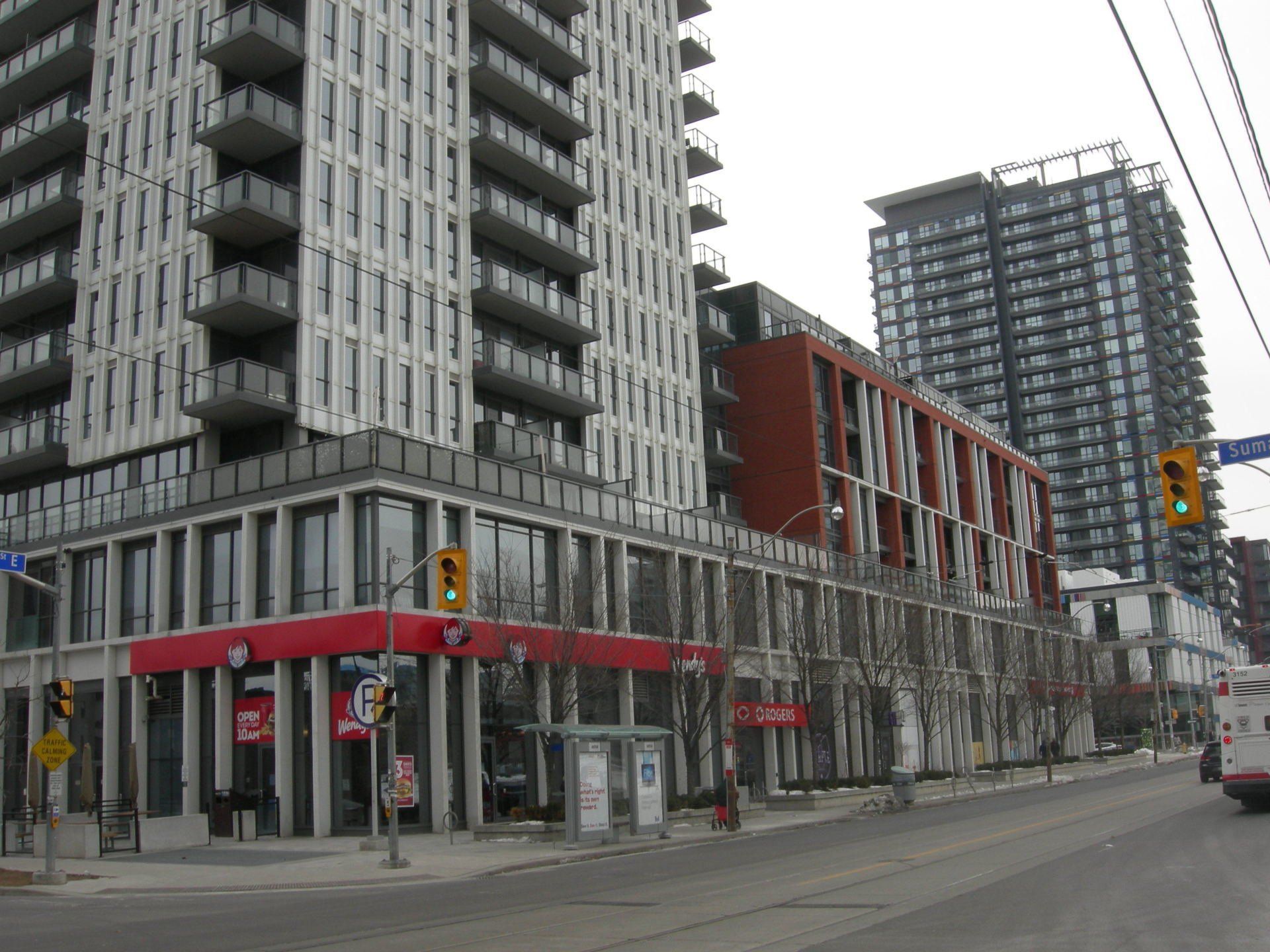 A street-level view of a modern white apartment building with a red-fronted business on the corner and a dark skyscraper.