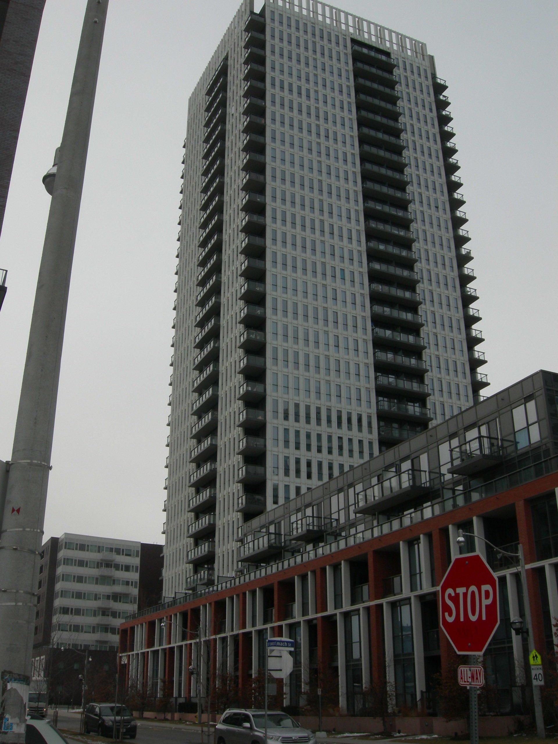 Tall, modern white residential tower with dark balconies above a brick podium, viewed from a street with a stop sign.