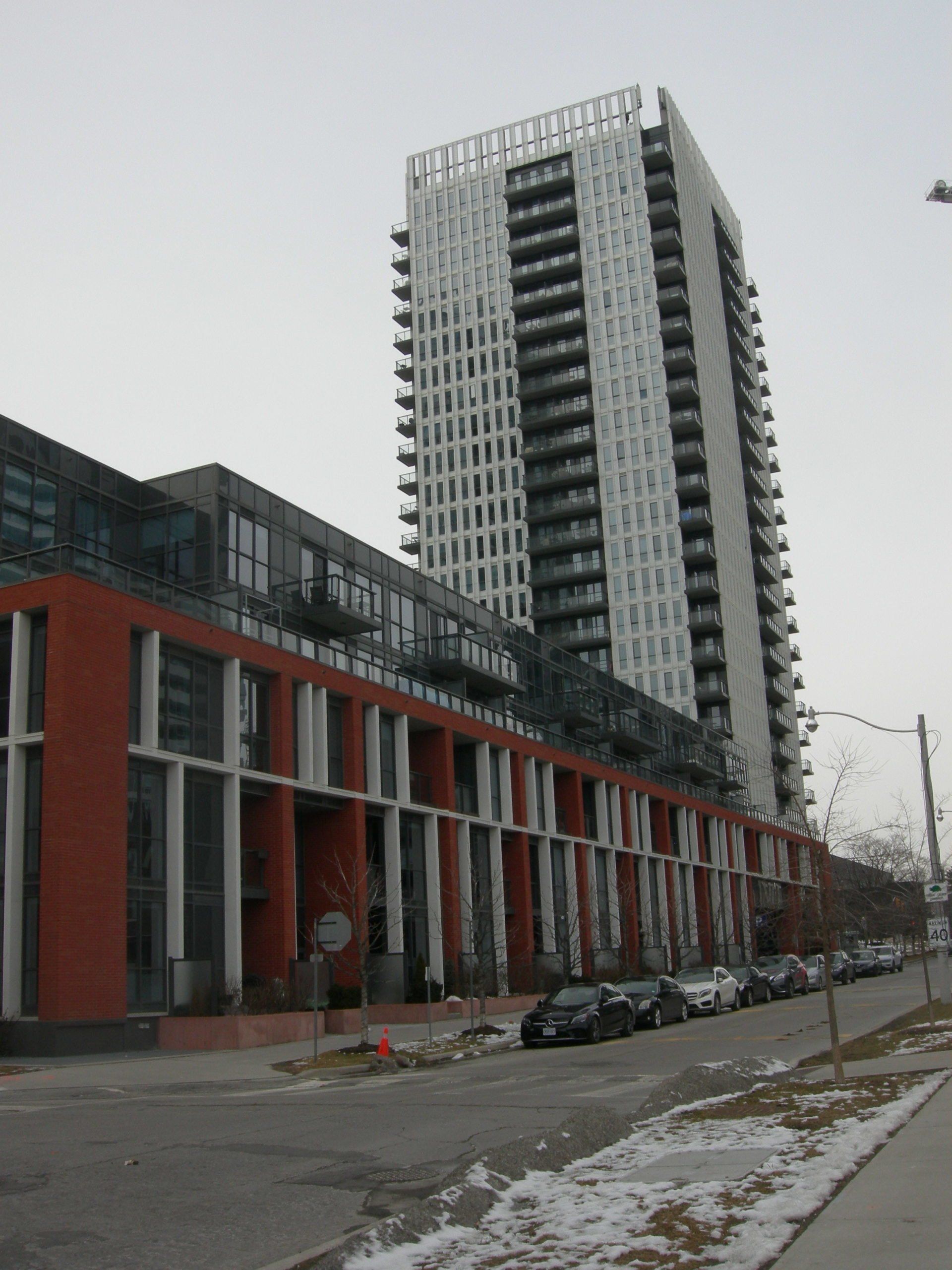 A tall, modern residential tower rises behind a multi-story, red-brick building lining a street with parked cars.