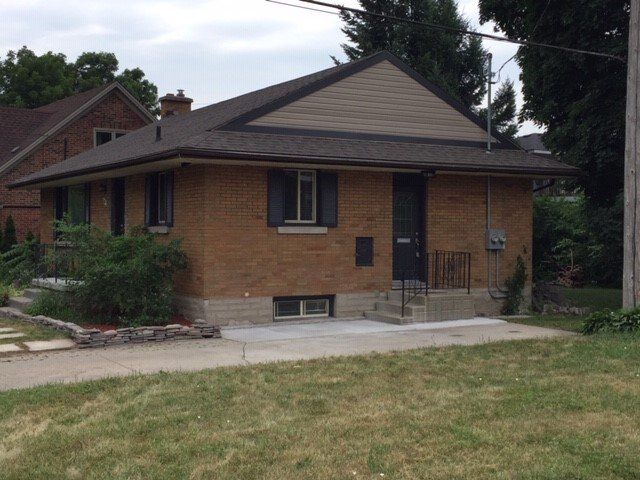 A single-story brown brick house with a dark roof and a small concrete entrance in a residential yard.